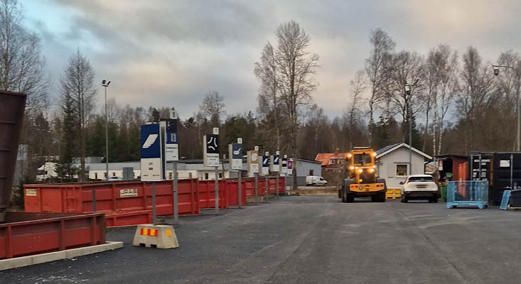 Översikt över Smålandsstenars återvinningscentral. Till vänster syns röda containrar med tydliga skyltar för varje fraktion. Till höger syns en hjullastare.