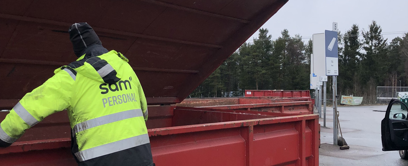 En person i varseljacka med SÅM-logotyp på ryggen står vid en röd container på en återvinningscentral.