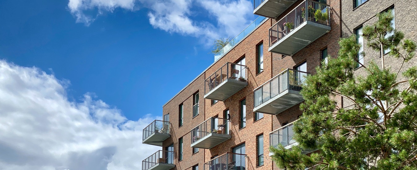 En bit av ett flerbostadshus i brunt tegel, med stora fönster och balkonger. I bakgrunden syns en blå himmel med stackmoln.