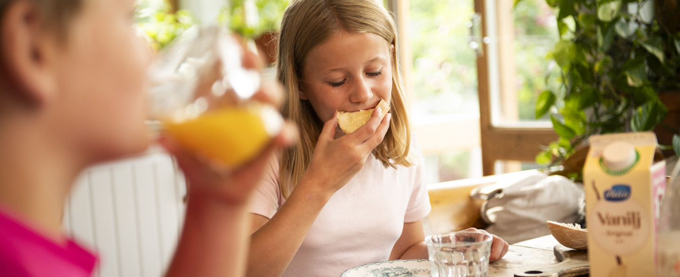 En ung person äter en macka. På bilden syns också en annan ung person som dricker ur ett glas med gul juice.