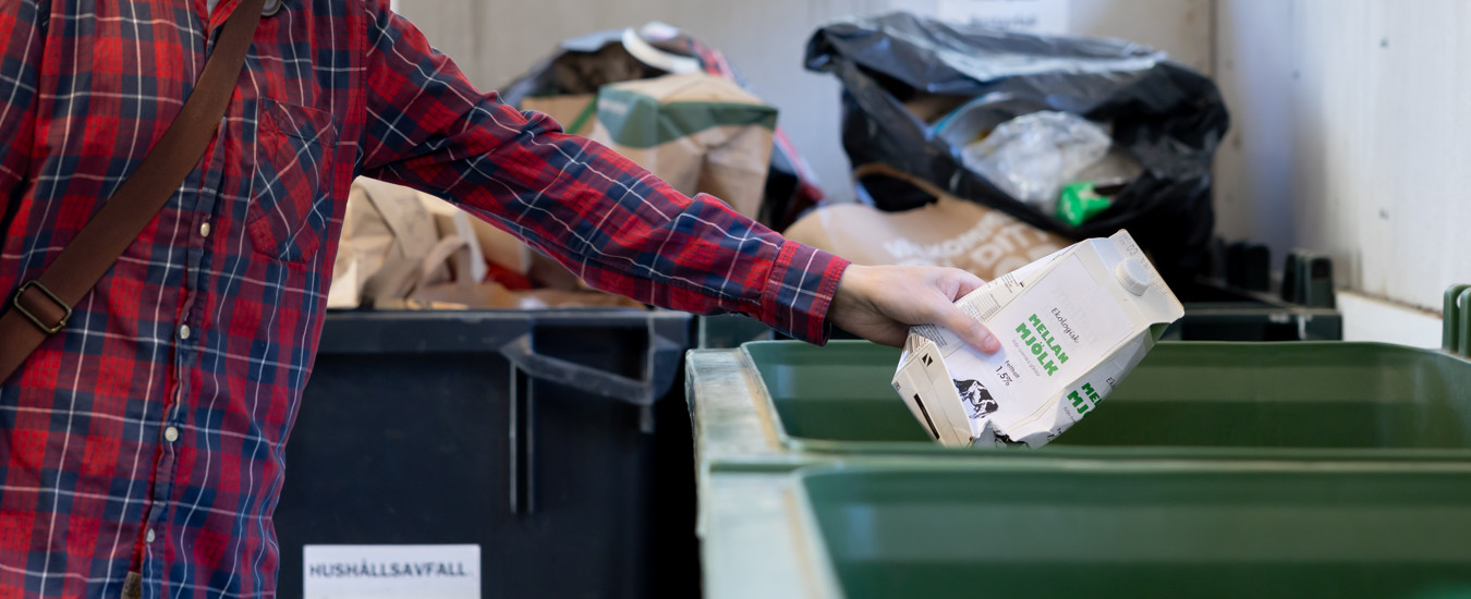 En person i rutig skjorta slänger en tom mjölkförpackning i ett kärl i ett miljöhus.