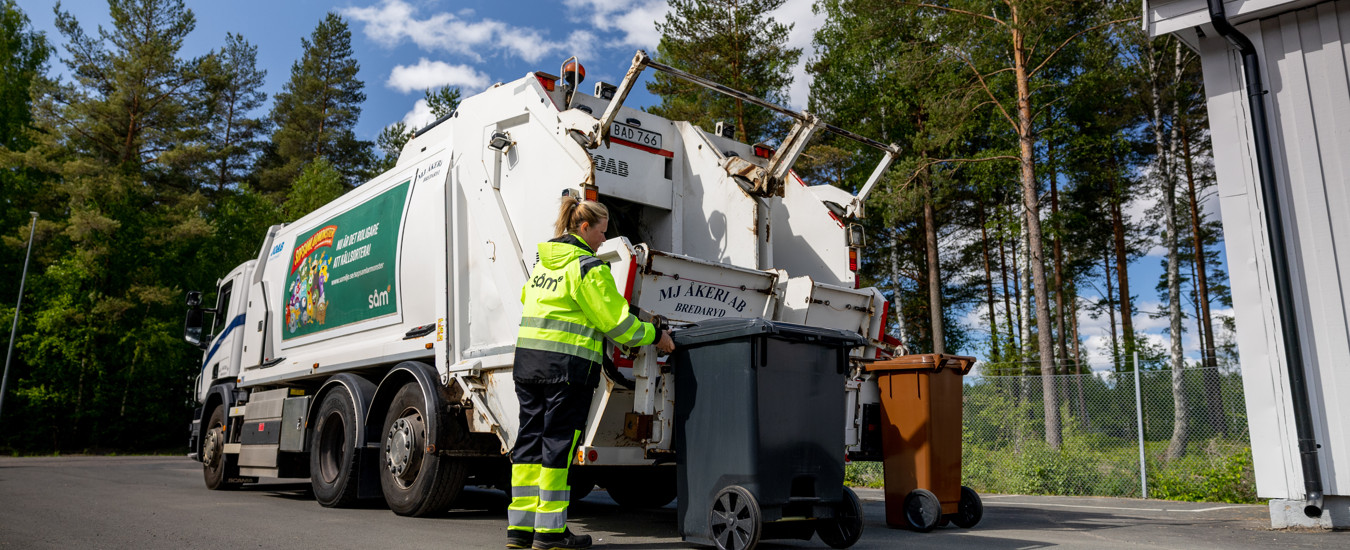 En sopbilschaufför i varselkläder i färd med att tömma två avfallskärl i en sopbil med två fack. 