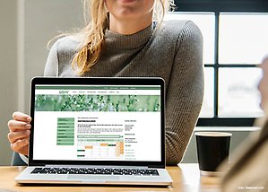 En person med grå tröja och blont hår visar upp en laptop med skärmen mot kameran. På laptopen syns SÅM:s hemsida med hämtningskalendern. 