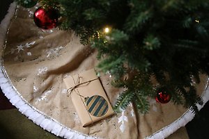 Ett paket under en julgran. Paketet är inslaget i brunt papper och har en målad julgranskula på ovansidan. Runtom paketet ligger barr från granen.