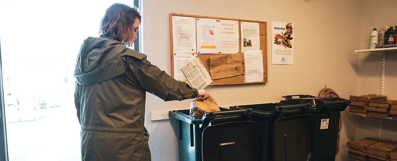 En person slänger en matavfallspåse i ett avfallskärl i ett miljöhus.
