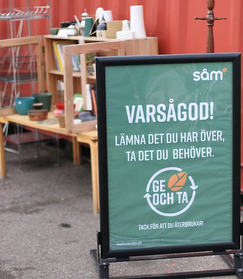 Skylt med texten Varsågod och information om Ge och Ta. I bakgrunden ett bord och ett skåp med Ge och Ta-grejer.