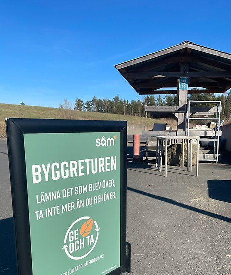 Skylt med texten Byggreturen och information om Ge och Ta. I bakgrunden en träställning med återbrukbart byggmaterial.