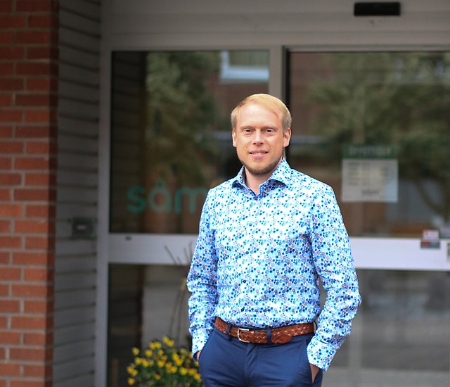 Porträtt av en leende person i blå blommig skjorta. Personen har ljust kort hår.