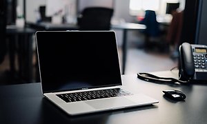 Dator och telefon på ett mörkt skrivbord i ett kontorslandskap.