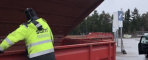 En person i varseljacka med SÅM-logotyp på ryggen står vid en röd container på en återvinningscentral.