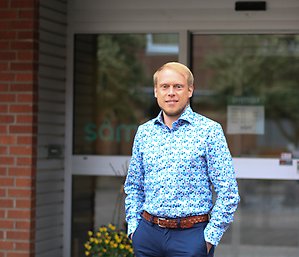 Porträtt av en leende person i blå blommig skjorta. Personen har ljust kort hår.