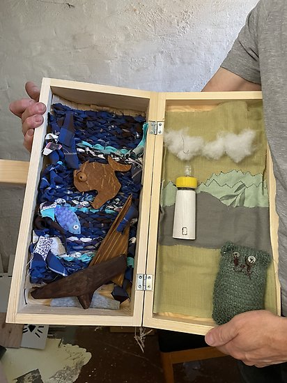 Ett litet träskåp med två dörrar, där den vänstra dörren representerar hav och den högra dörren representerar land. Inredningen är en kombination av vävd, virkad och hopsydd textil samt träfigurer.