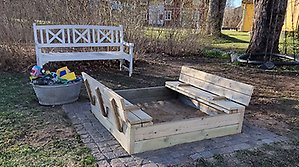 Färdigbyggd sandlåda med ett lock som fälls upp till en bänk på var sida av sandlådan.